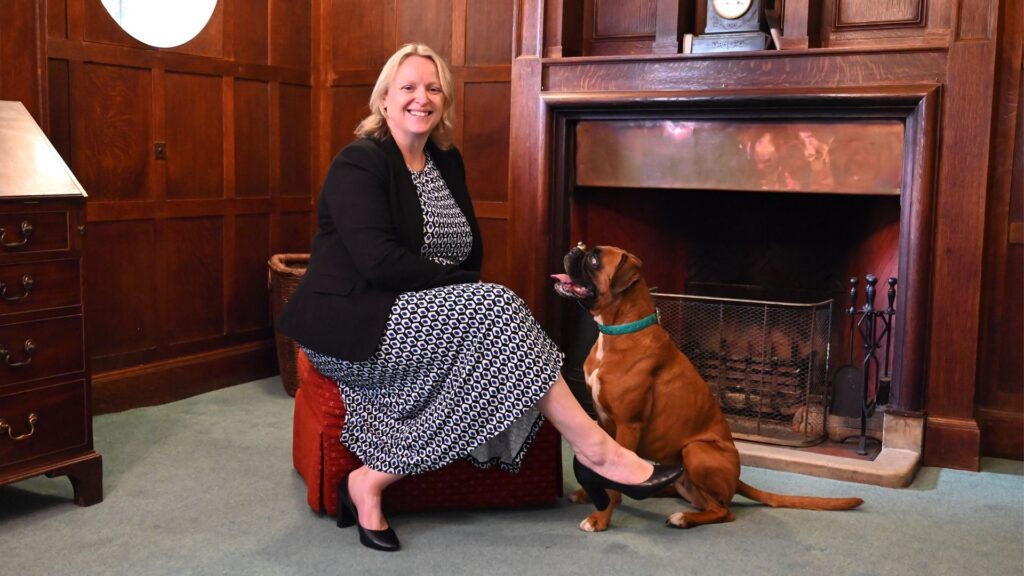 Claire Gilding-Brant, Interim Head of Luckley House School, sitting in front of a fireplace with a dog at her feet, smiling warmly.